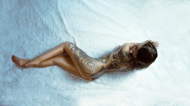 Krásna nahá žena s potetovaným telom leží na bielej deke.jpg